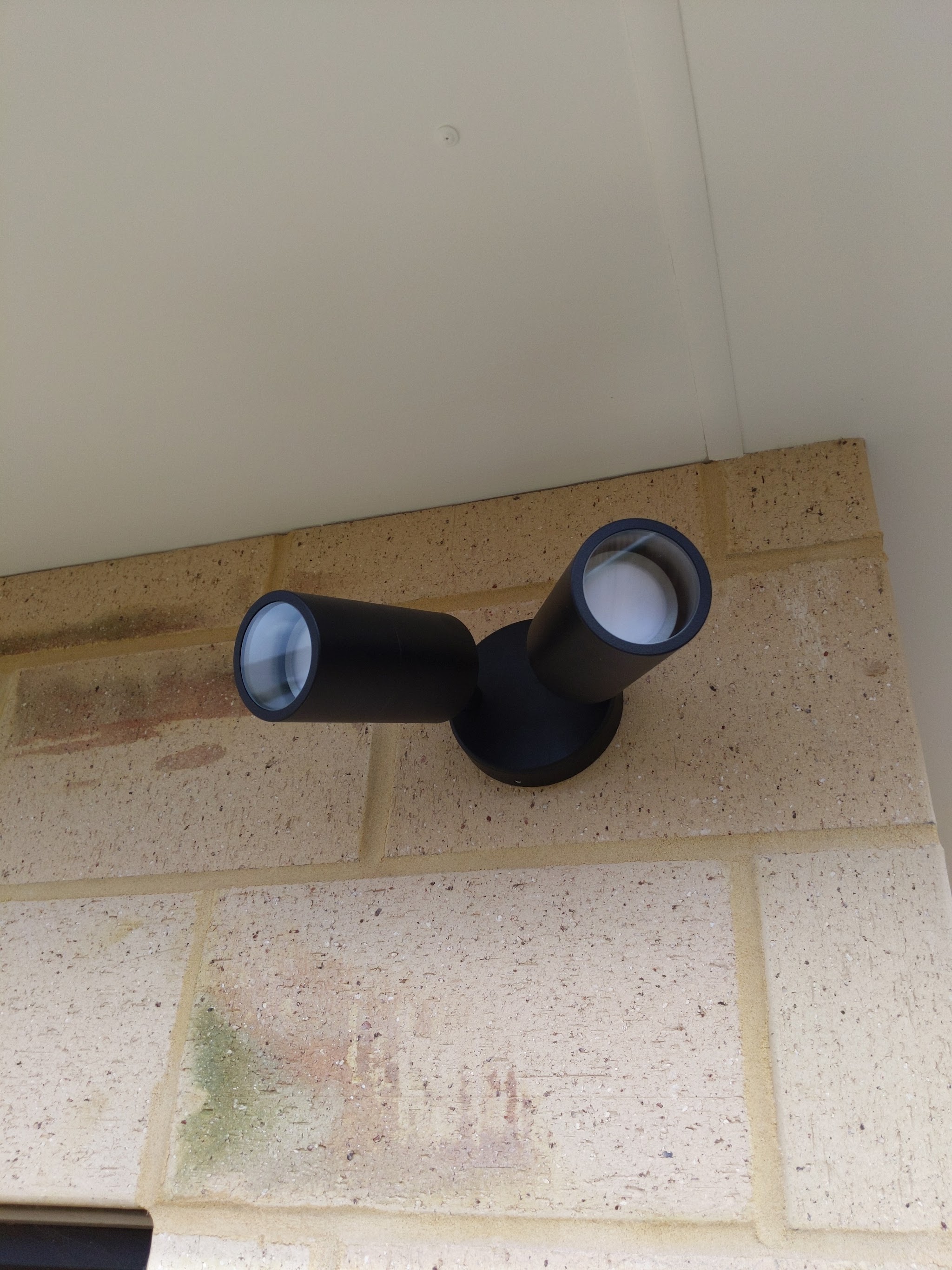 Black twin-head outdoor spotlight installed under a brick eave